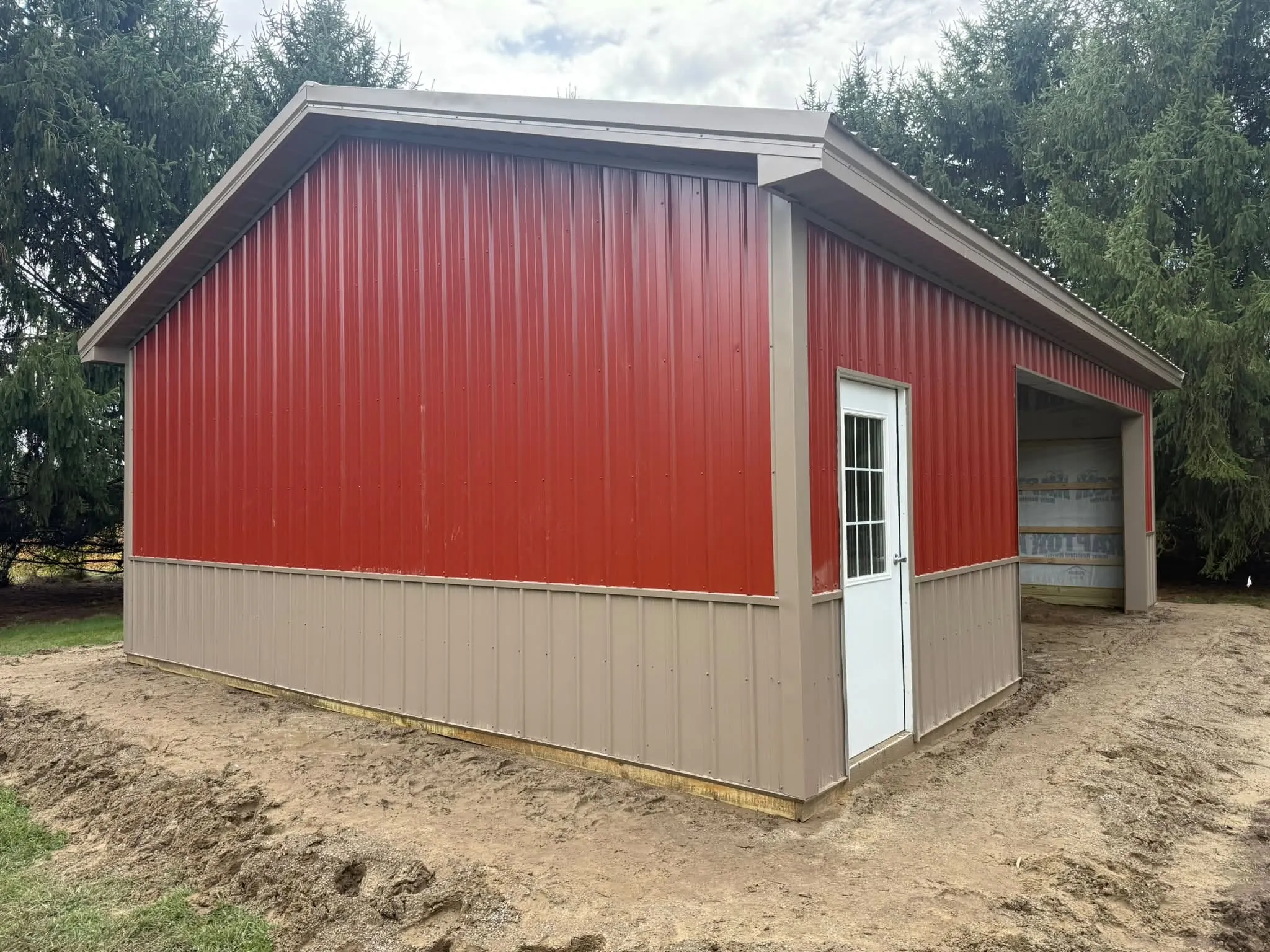 The finished pole barn in red steel with a tan wainscot and tan trim, a white walk door with a window, and an open bay on the side