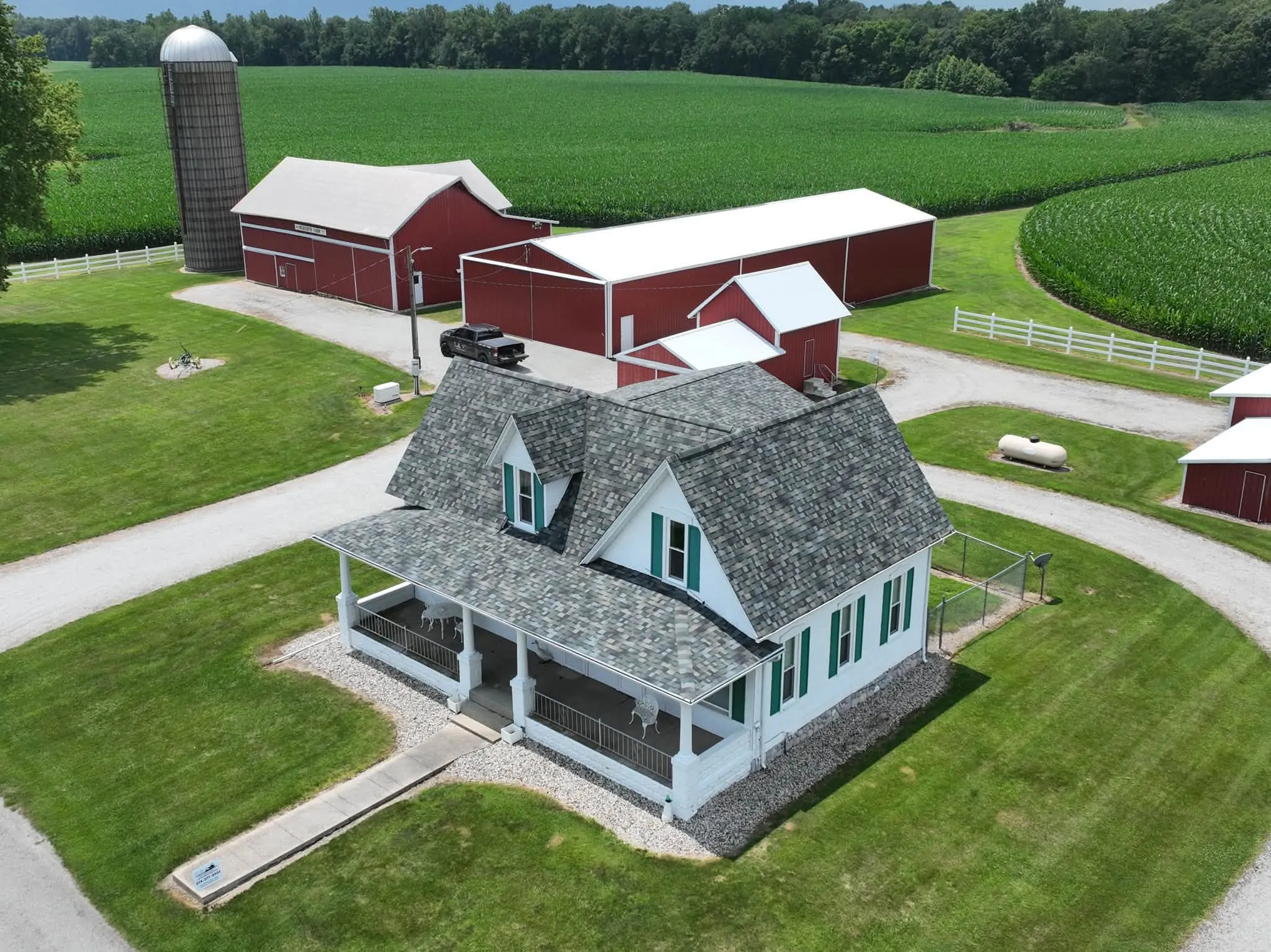 The finished farmhouse from the air, with its new Evergreen Mist shingle roof and the farm's red barns and silo behind it