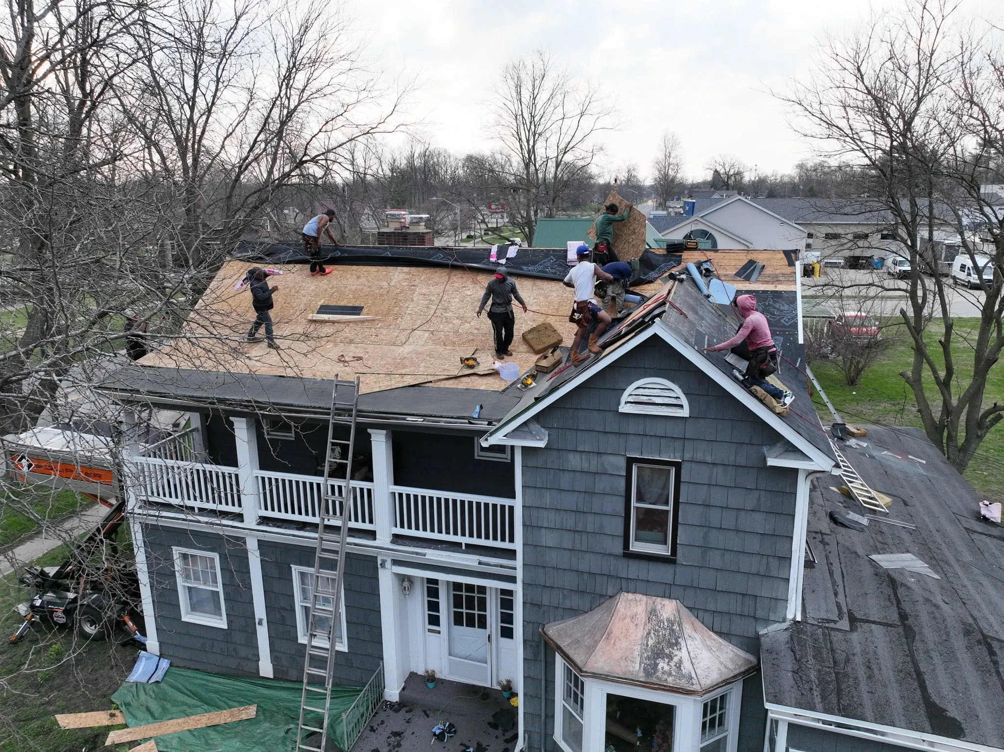 The crew laying new decking across the low-slope section of the roof, with the two-tier front porch and the town beyond