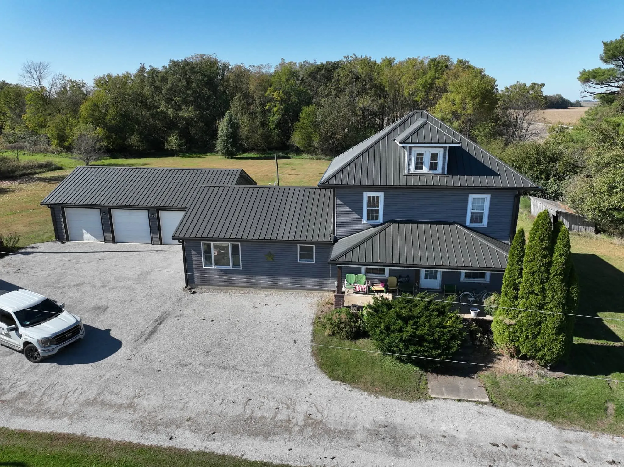 The finished farmhouse from above the drive, with the new Dark Slate metal roof across the two-story house, the porch roofs, and the long wing to the garage