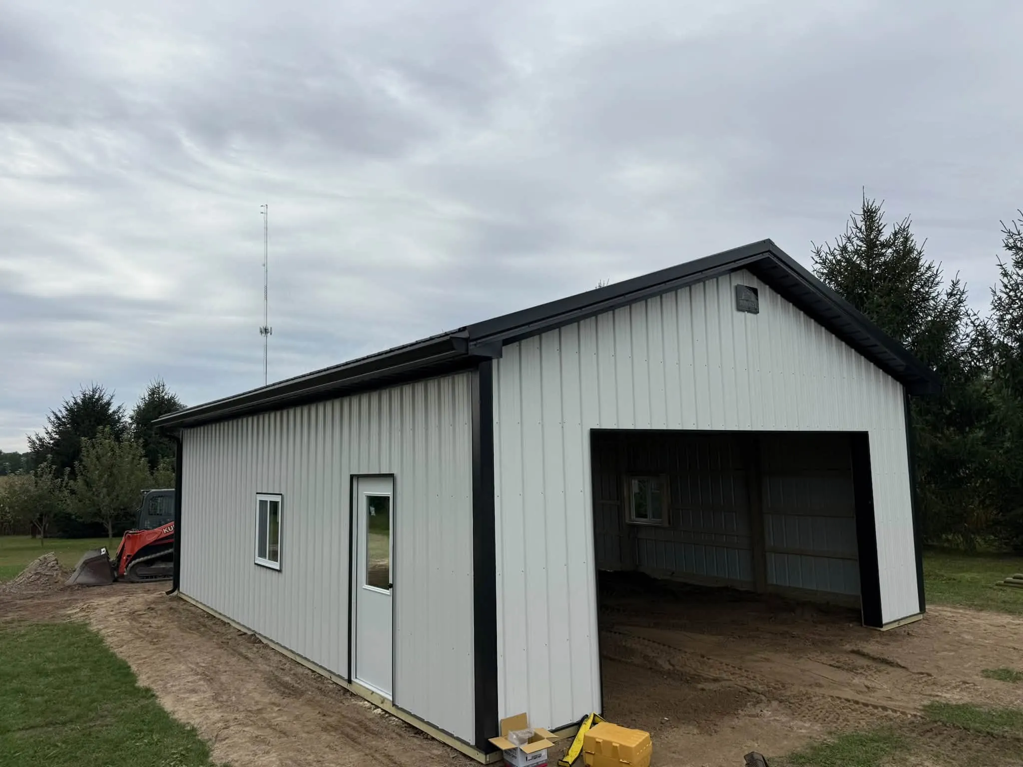 The finished pole barn in white steel with black trim, with a walk door and a window along the side and a large opening at the gable end