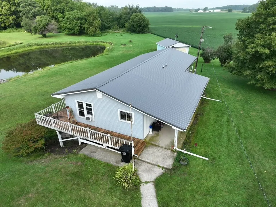 The finished metal roof from above, on a ranch home with a raised deck, a pond, and farm fields beyond