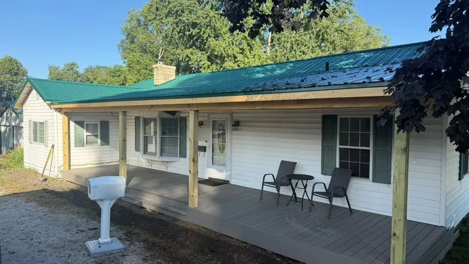 The finished house with its new green metal roof carried out over the new front deck on wood posts, with two chairs and a small table on the deck