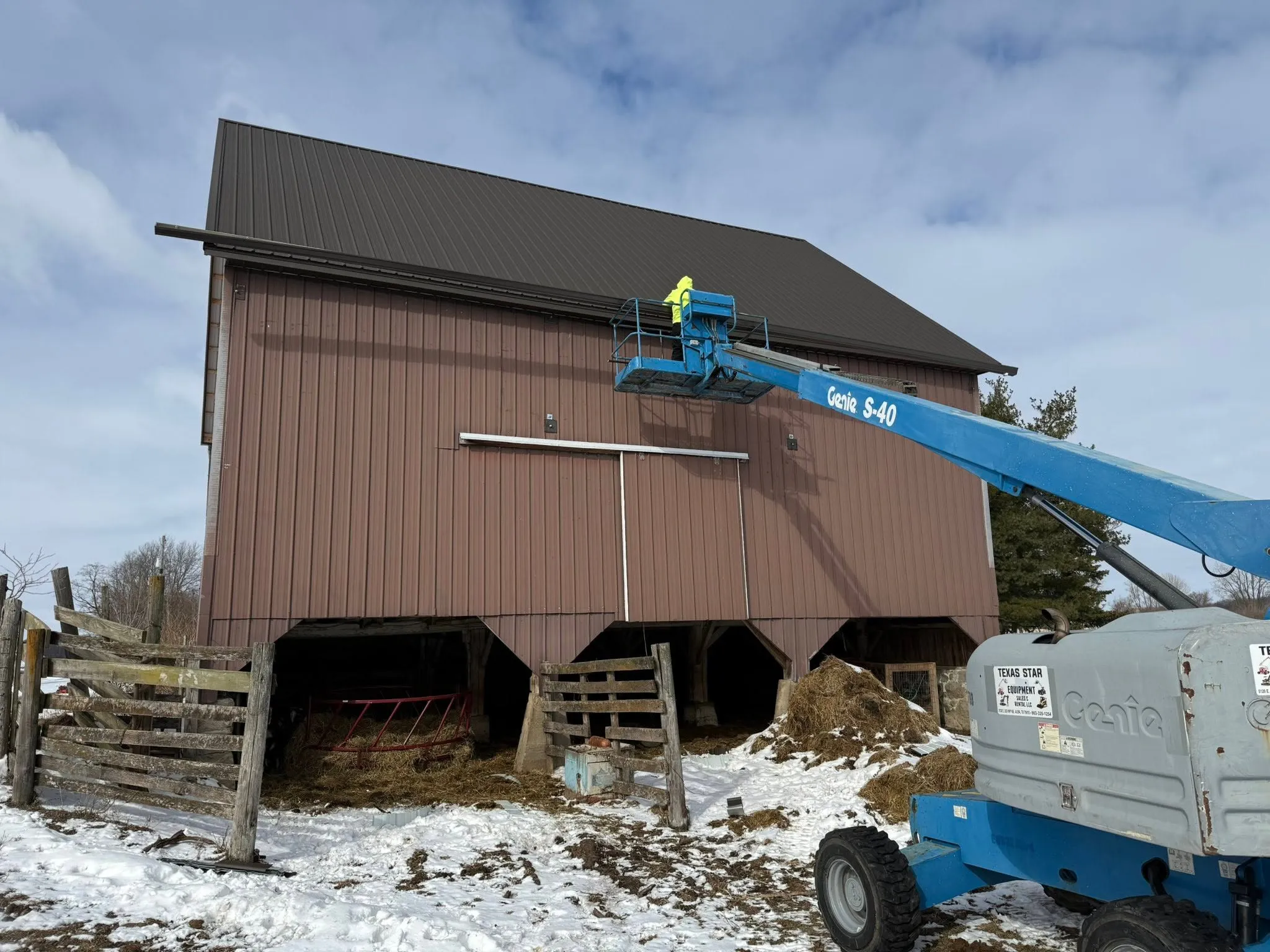 A worker in a boom lift fitting gutter along the eave of a large brown barn, with snow on the ground