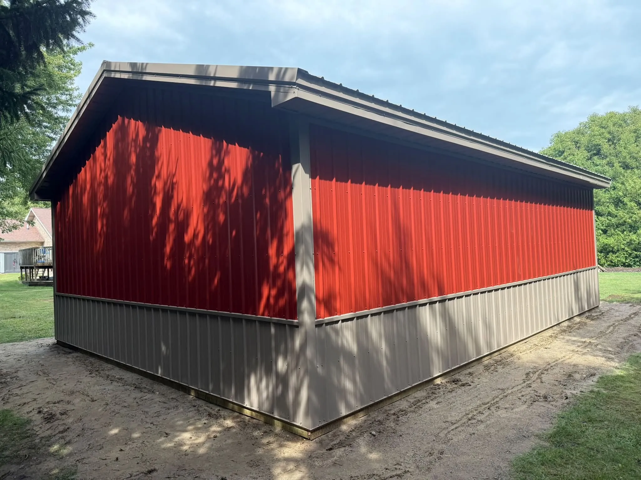 The long side of the barn in red steel over the tan wainscot, with evergreens and a house in the background