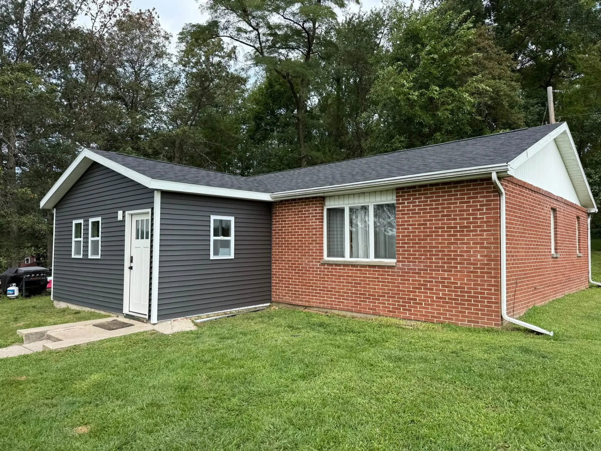 The finished addition from the yard, with dark gray siding, white trim, a white door and two windows, joined to the brick house