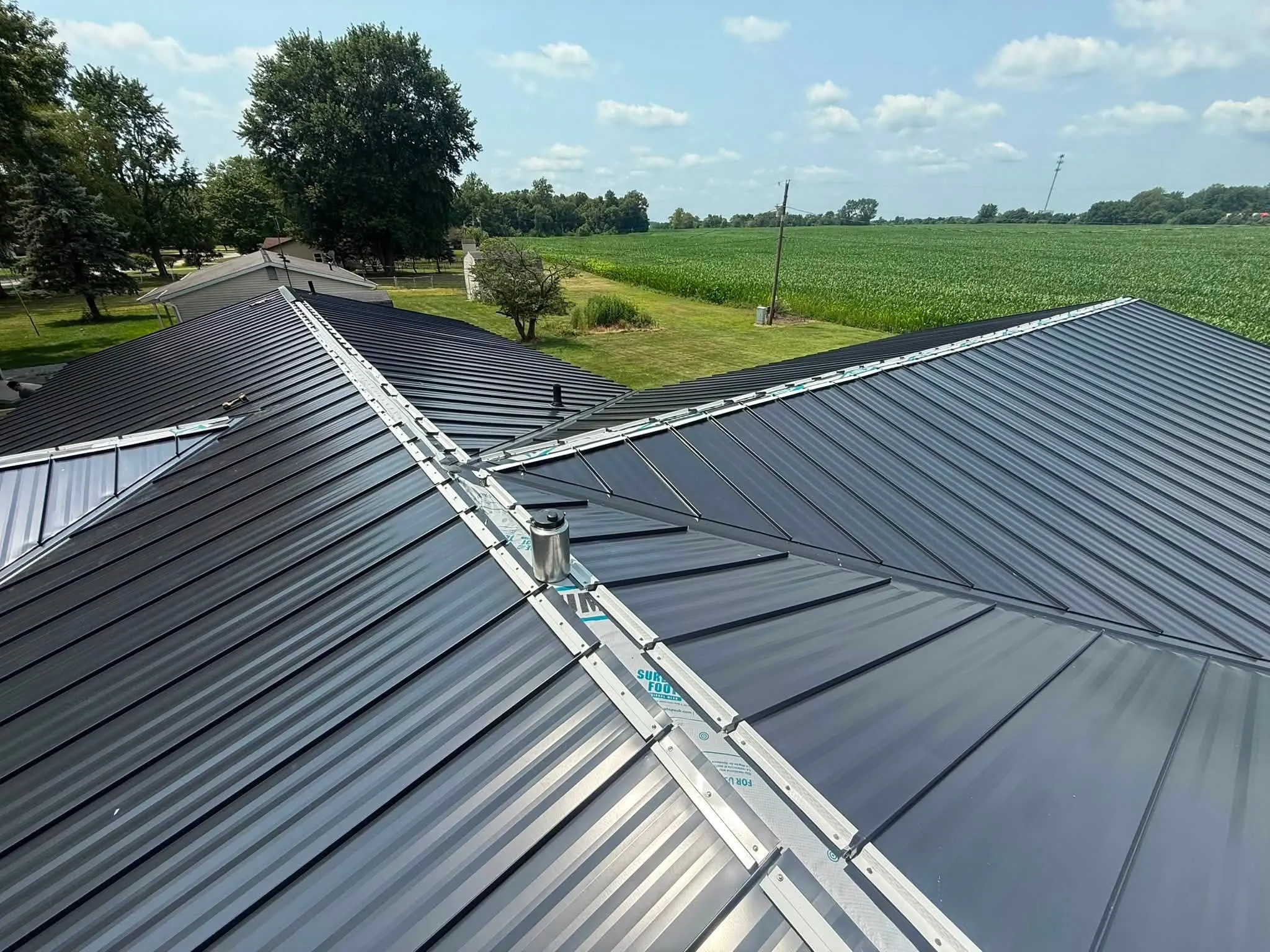 Standing seam panels running up the slope to the hip, with the ridge trim not yet fitted and a cornfield beyond