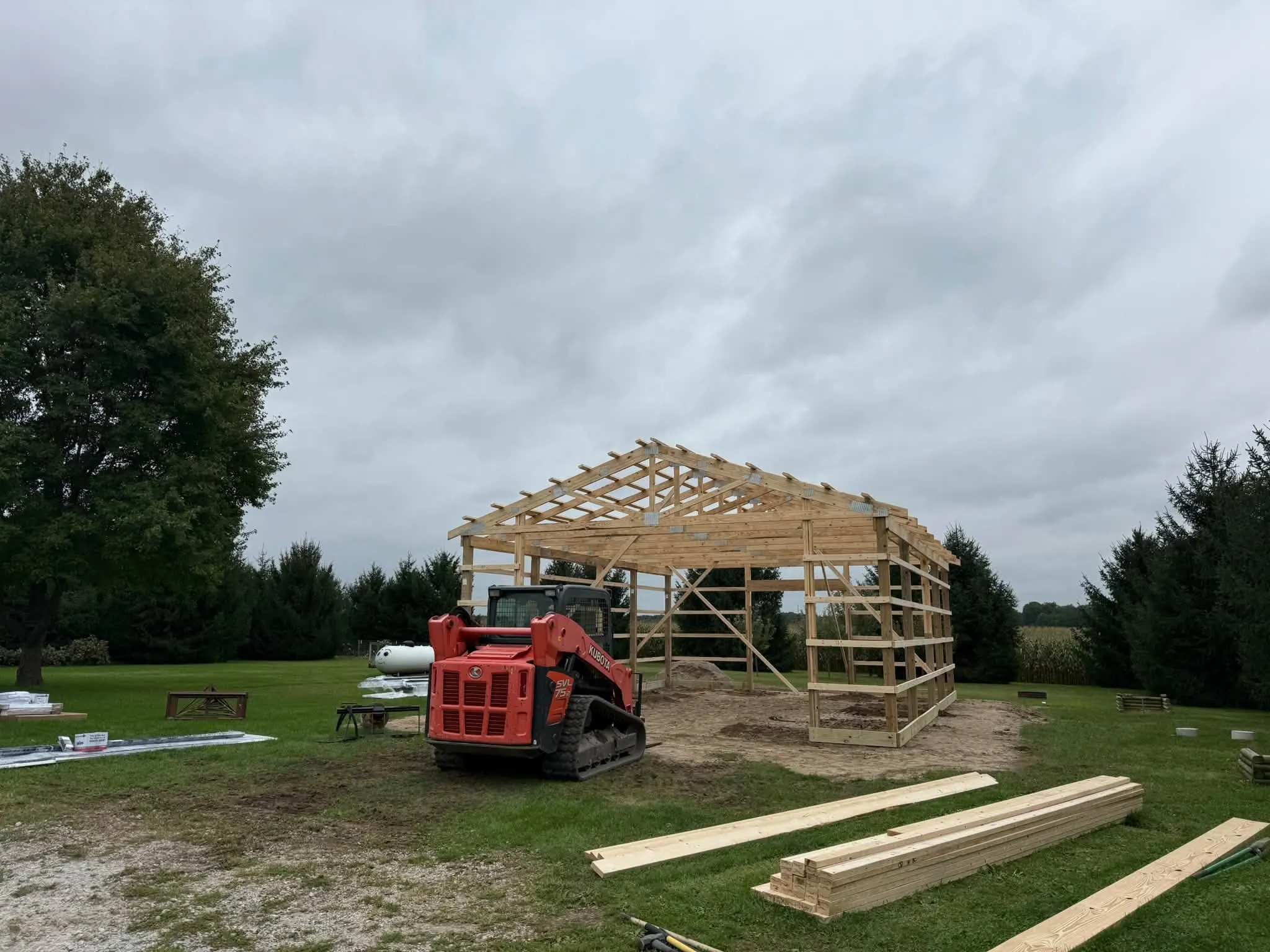 The wood frame and trusses standing in the yard, with a tracked skid steer beside it and lumber laid out on the grass