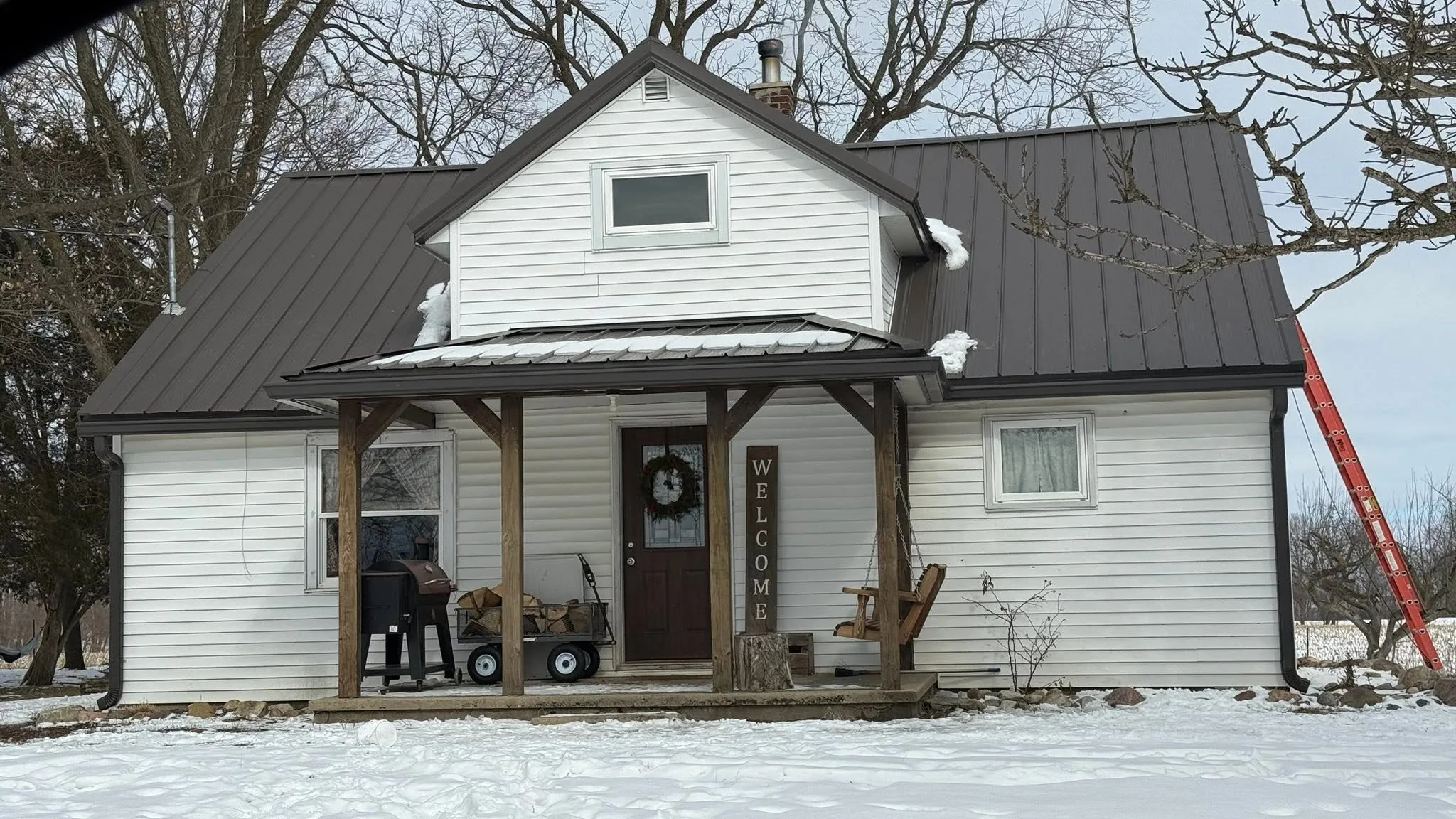 The white farmhouse with its new brown gutters and downspout, timber porch, and snow across the yard