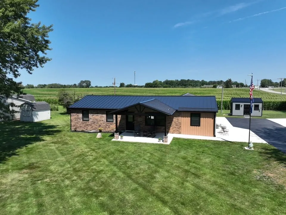 The finished brick ranch under its new dark standing seam roof, with the gabled front porch, a flagpole, and cornfields beyond