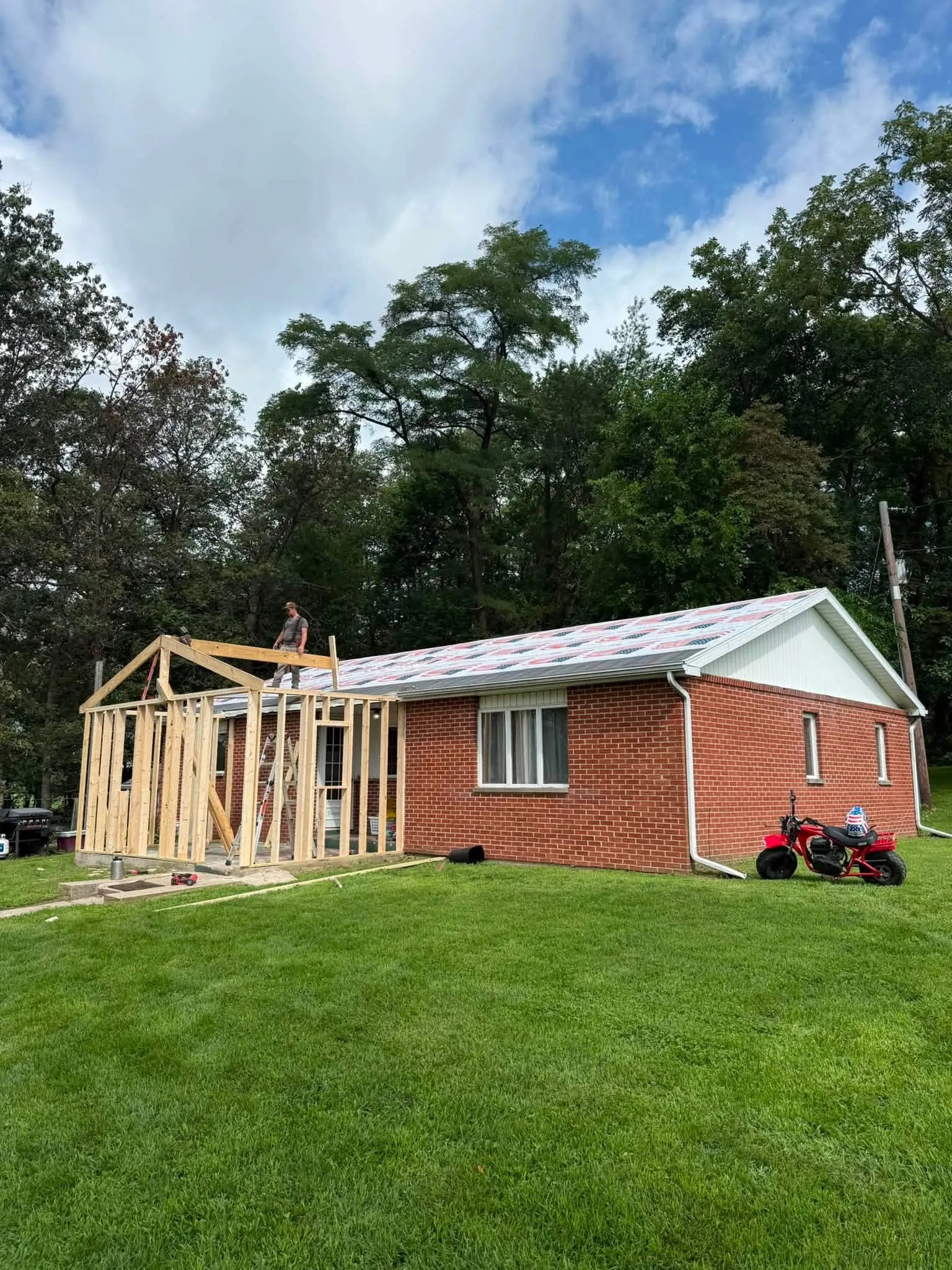The addition framed in bare wood against the end of the brick house, with a crew member up on the frame and new underlayment on the main roof