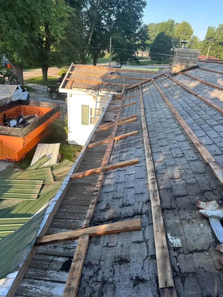 The tear-off in progress, with the old green panels piled by the dumpster and the old shingles and wood strips still on the roof