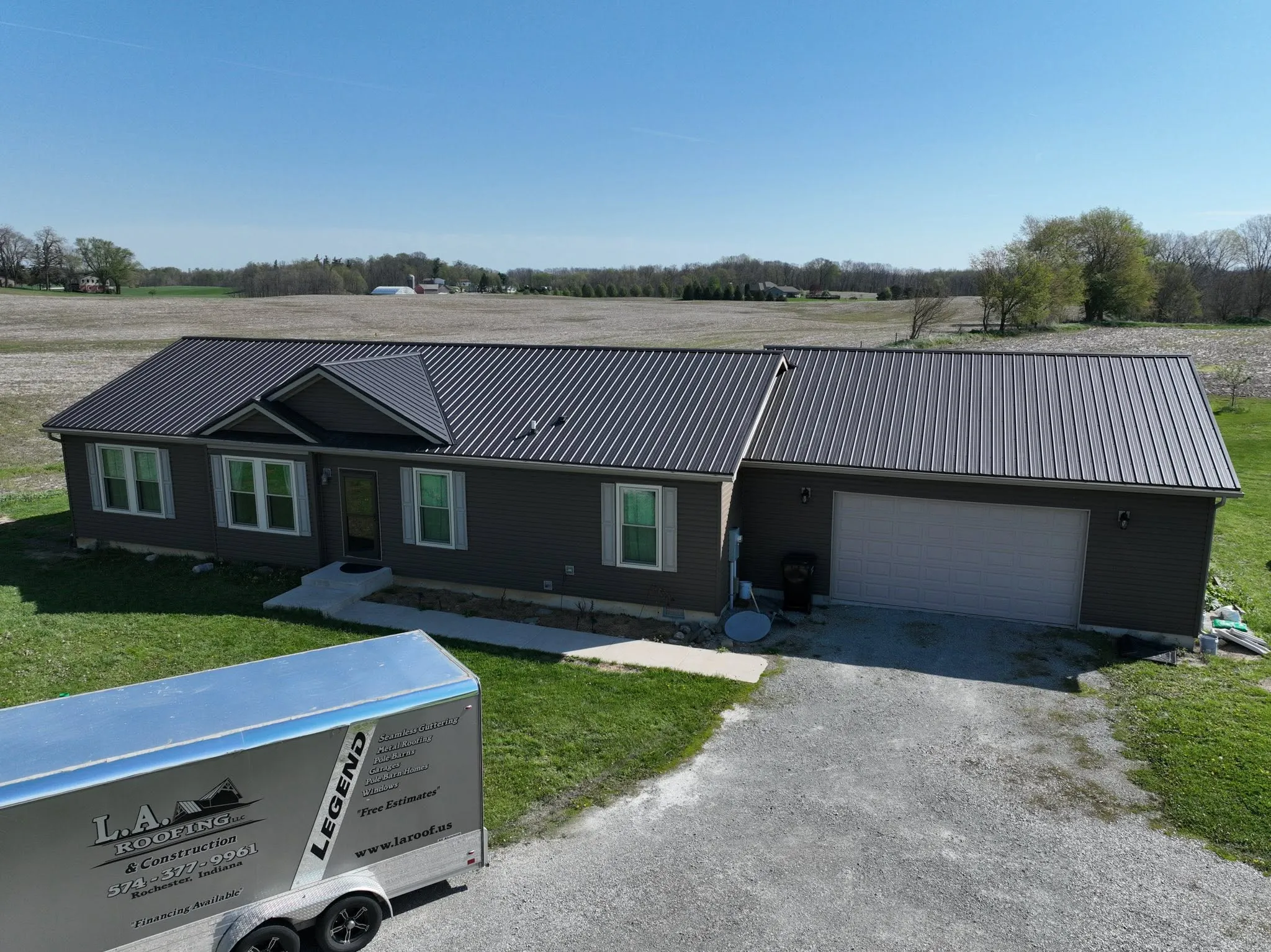 The completed roof from the driveway, with the L.A. Roofing trailer parked in front and farm fields behind