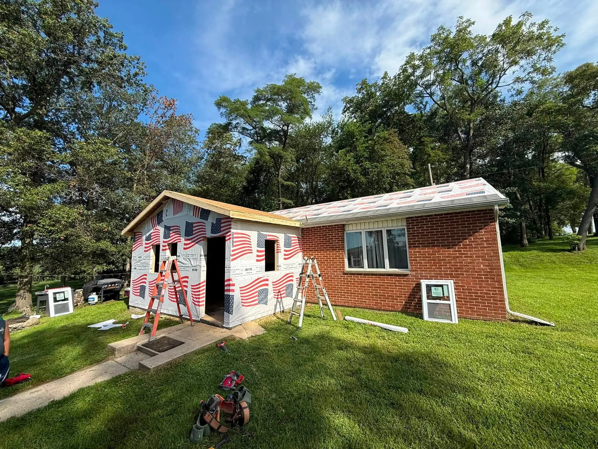 The addition sheathed and covered in flag-printed housewrap, with ladders set up and the doorway still open