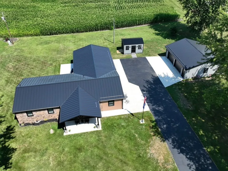 High view over the front of the house, showing the hips and the porch gable in standing seam, with the detached garage and a shed beside the drive